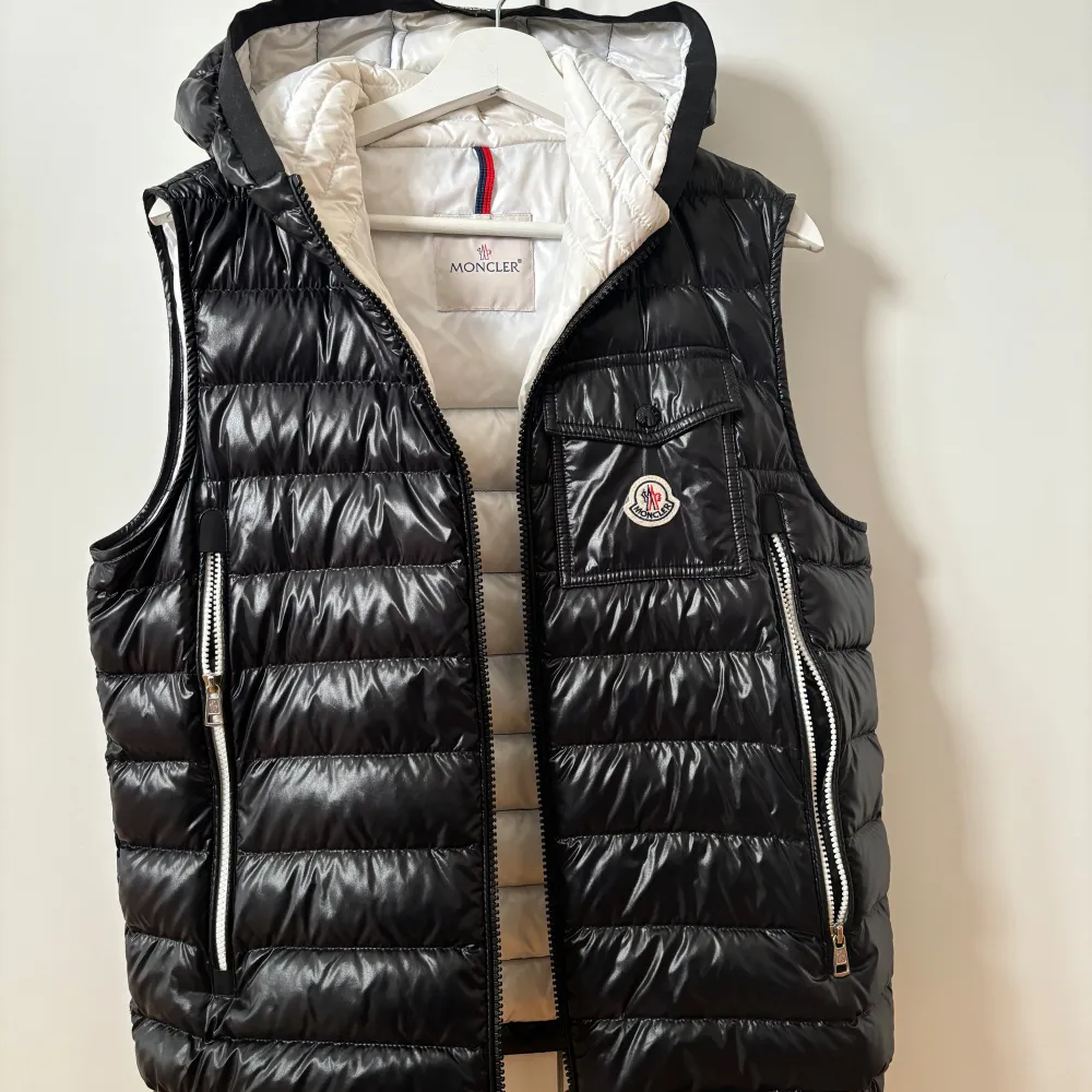 Säljer en snygg svart dunväst från Moncler. Västen har en quiltad design med dragkedja framtill och två praktiska fickor med dragkedjor. Den har också en liten bröstficka med Moncler-loggan. Insidan är vit och västen har en avtagbar huva. Perfekt för kyliga höst- och vinterdagar!. Jackor.