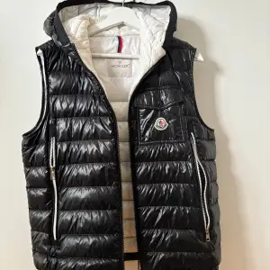 Säljer en snygg svart dunväst från Moncler. Västen har en quiltad design med dragkedja framtill och två praktiska fickor med dragkedjor. Den har också en liten bröstficka med Moncler-loggan. Insidan är vit och västen har en avtagbar huva. Perfekt för kyliga höst- och vinterdagar!