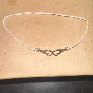 Säljer ett enkelt och stilrent armband med en silverfärgad vinge i mitten. Armbandet har en tunn vit tråd som gör det justerbart och bekvämt att bära. Perfekt för vardagsbruk eller som en fin detalj till en festoutfit.