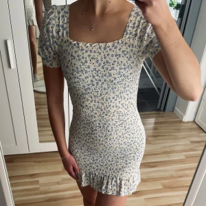 Blommig kortklänning med volang - Säljer en superfin blommig kortklänning med volangdetaljer. Klänningen har en tight passform och korta ärmar. Den är cremevit med små blommor i blått.