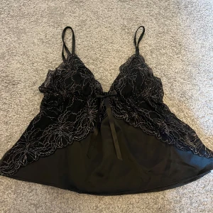 Svart spetslinne med rosett - Säljer ett elegant svart spetslinne med tunna axelband och en söt rosett framtill. Linnet har vackra spetsdetaljer och en lätt transparent look, perfekt för en festlig kväll. Det är i nyskick och passar perfekt för en chic och stilren outfit.