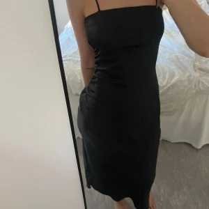 Svart axelbandslös klänning - Snygg svart klänning med smala axelband och en elegant slits. Perfekt för festliga tillfällen. Klänningen har en tight passform i satin🖤🖤