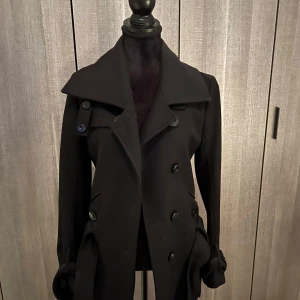 Svart jacka med knappar - Säljer en elegant svart jacka med dubbelknäppning och stora knappar. Den har en klassisk krage och långa ärmar, perfekt för höst och vinter. Passar både till vardags och festliga tillfällen. 