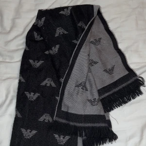 Svart och grå halsduk från Emporio Armani - Snygg och stilren halsduk från Emporio Armani i svart och grått. Den har ett diskret mönster med märkets logotyp och fransar i ändarna. Perfekt för att hålla sig varm och trendig under kyliga dagar.