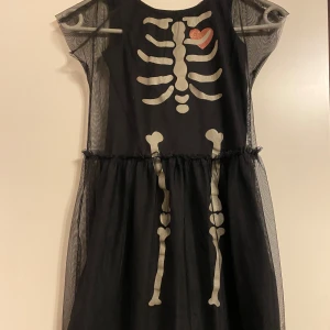Svart skelettklänning från H&M  - Cool svart klänning från H&M med skelettmönster och ett rött hjärta på bröstet. Skelettet är glow in the dark. Perfekt för Halloween eller maskerad! Klänningen har korta ärmar och ett tunt tyllöverdrag som ger en extra dimension. Passar barn i storlek 122.