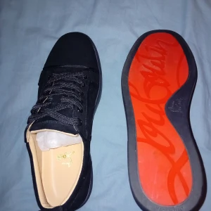 Svarta sneakers med röd sula - Säljer ett par snygga svarta sneakers från Christian Louboutin. De har en ikonisk röd sula och är i mocka med snörning. Perfekta för både vardag och fest! Skorna är i nyskick och har en stilren design som passar till det mesta.