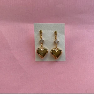Guldiga hjärtformade örhängen - Säljer ett par handgjorda örhängen i guld med hjärtformad design. De är 3,5 cm långa och 1,5 cm breda, perfekta för att ge en romantisk touch till din outfit. Örhängena har en liten glittrande detalj ovanför hjärtat som ger extra bling.