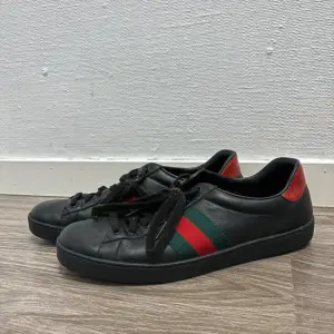 Säljer ett par svarta Gucci sneakers i skinn med klassiska gröna och röda ränder på sidan. Skorna har snygga röda detaljer på hälen och är i bra skick. Perfekta för både vardag och fest! 