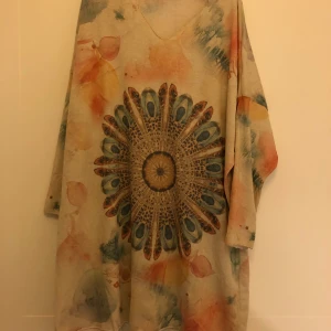 Färgglad lång tunika med mandalamönster - Säljer en unik och färgglad lång tunika med ett stort mandalamönster i mitten. Klänningen har långa ärmar och ett löst passande snitt, perfekt för en bohemisk stil. Materialet känns mjukt och bekvämt, och färgerna är en mix av beige, blå, orange och rosa. Perfekt för höstfest eller en avslappnad dag på stan!