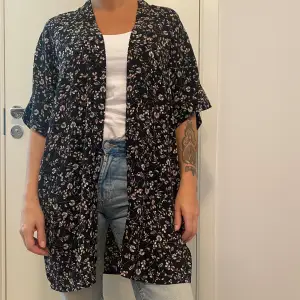 Säljer en superfin svart kimono från H&M med blommigt mönster i ljusa färger. Den har korta ärmar och en lös passform, perfekt för vår och sommar. Använd den över en enkel topp för en stilren look. 🌸