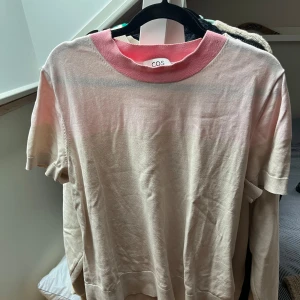 Beige och rosa tröja från COS - Säljer en snygg beige tröja från COS med rosa detaljer vid halsringningen. Tröjan är kortärmad och perfekt för vår och sommar. Den är i bra skick och passar både till vardags och lite finare tillfällen. materialet är lite tjockare än vanligt T-shirtmaterial. 