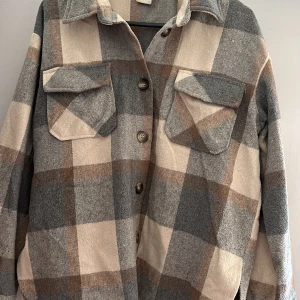Rutig overshirt i beige och grå - Säljer en superfin rutig overshirt i beige och grå toner. Den har en klassisk krage och knappar framtill. Två bröstfickor med lock ger en cool look. Perfekt för höst och vår!