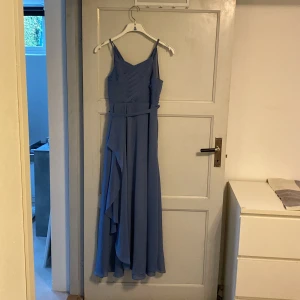 Blå långklänning med volangdetalj - Säljer en elegant blå långklänning med smala axelband och en vacker volangdetalj på sidan. Perfekt för sommarens fester eller en bal. Klänningen har en figursmickrande passform och är tillverkad i ett lätt och luftigt material. priset kan diskuteras