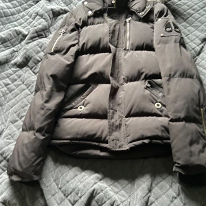 Svart pufferjacka från Moose Knuckles - Säljer en svart pufferjacka från Moose Knuckles. Jackan är fylld med dun och har en mysig pälsdetalj vid kragen. Den har flera fickor med dragkedjor och knappar, perfekt för att hålla värmen under kalla vinterdagar. Jackan är i bra skick och redo för nya äventyr, förutom att märeket på jackan har blivit av med lite färg, BARA SERIÖSA❤️