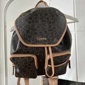 Brun ryggsäck från Calvin Klein - Säljer en snygg brun ryggsäck från Calvin Klein med deras logotypmönster. Väskan har flera praktiska fack med dragkedjor och justerbara axelremmar med märkesdetaljer. Perfekt för både vardag och utflykter!