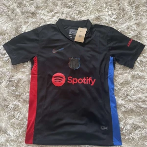 Svart fotbollströja från Nike - Säljer en svart fotbollströja från Nike med korta ärmar. Tröjan har röda och blå detaljer på sidorna och ett Spotify-tryck på framsidan. På baksidan finns UNHCR-loggan. Perfekt för fotbollsfans som vill ha något unikt!