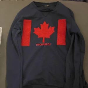 Mörkblå tröja från DSQUARED2 - Säljer en snygg mörkblå tröja från DSQUARED2 med ett rött lönnlöv och text på framsidan. Tröjan har långa ärmar och en klassisk passform. Perfekt för dig som vill ha något stilrent och bekvämt. Hör av dig vid intresse! Priset kan diskuteras vid snabbt affär 