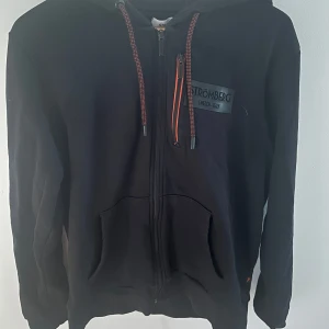 Svart hoodie från Strömberg - Säljer en svart hoodie från Strömberg i bomull. Den har dragkedja framtill och en praktisk huva med snören. Perfekt för en avslappnad stil.