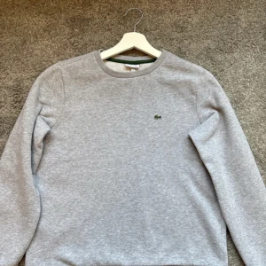 Grå sweatshirt från Lacoste - Säljer en stilren grå sweatshirt från Lacoste med det klassiska krokodilemblem på bröstet. Tröjan har långa ärmar och en rund halsringning med en grön detalj på insidan av kragen. Perfekt för en avslappnad look. använd ett fåtal gånger och är i topp kvalité. storlek s eller 176cm