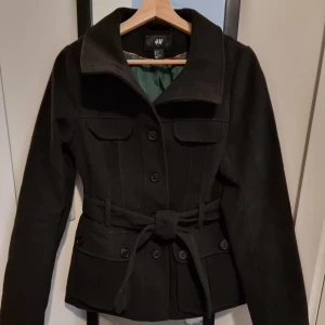 Svart kappa från H&M - Snygg svart kappa från H&M med knappar och bälte i midjan. Jackan har en klassisk krage och två fickor framtill. Perfekt för kyligare dagar med sin stilrena design.