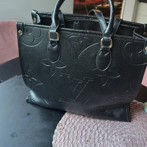 Svart axelväska med röd insida - Snygg svart axelväska i skinn med elegant präglat mönster. Väskan har en rymlig röd insida och praktiska handtag. Perfekt för att bära med sig allt man behöver med stil.