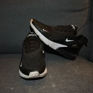 Svarta Nike Air Max 270 - Snygga svarta Nike Air Max 270 sneakers med vit sula och ikonisk Nike-logga. Skorna har en luftig design med snörning och en synlig Air-enhet i hälen för extra komfort. Perfekta för en sportig look.