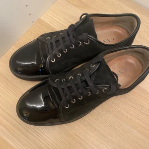 Helsvarta Lanvins med glansig cap toe - Snygga svarta lanvins med glänsande lackdetaljer på tån. Skorna har snörning och en stilren design som passar perfekt för en laidback stil. Tillverkade i en kombination av mocka och syntetmaterial. Skorna är i gott skick endast använt några gånger, säljs på grund av fel storlek. Skorna är i uk 12 EU 45-46. Har tyvärr ingen box men har lanvins tygpåse. Pris kan diskuteras!