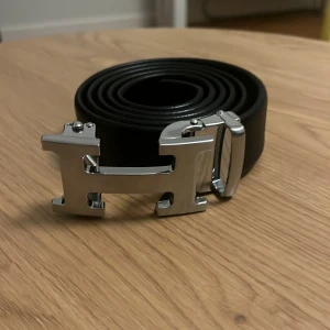 Svart bälte med H-spänne - Snyggt svart bälte med ett iögonfallande H-format spänne i silver. Perfekt för att ge en stilren touch till din outfit. Bältet är tillverkat i skinn och har en klassisk design som passar till många olika stilar. 