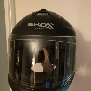 Svart moped hjälp från Shox - Säljer en svart hjälm från Shox med visir. Hjälmen har en stilren design med märkets logotyp på framsidan och baksidan. Perfekt för motorcykelentusiaster som söker både stil och säkerhet.