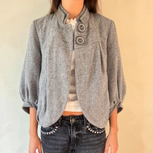 Grå kavaj med knappar - Snygg grå kavaj med trekvartsärm och två stora knappar framtill. Den har en elegant och stilren design med en lätt rundad nederkant. Perfekt för en chic look.