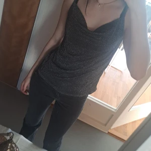 Glittrig svart topp - Snygg glittrig svart topp med draperad halslinje. Perfekt för en kväll ute eller en fest. Toppen har en smickrande passform och är ärmlös, vilket gör den både elegant och bekväm.