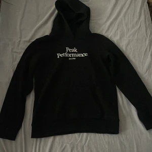 Svart hoodie från Peak Performance - Säljer en stilren svart hoodie från Peak Performance med vit logga på framsidan. Tröjan har en klassisk design med huva och långa ärmar. Perfekt för en avslappnad look.