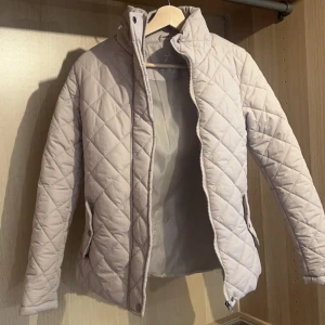 Beige quiltad vårjacka  - Snygg beige quiltad vårjacka med dragkedja och knappar. Perfekt för kyligare dagar med sin stilrena design och praktiska fickor. Jackan har långa ärmar och en bekväm passform. Finns en liten fläck (se bild) 