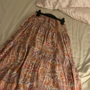 Mönstrad långkjol i bomull - Säljer en färgglad långkjol med ett vackert paisleymönster i rosa, orange och lila toner. Kjolen är gjord i ett lätt och luftigt bomullsmaterial, perfekt för varma dagar. Den har en elastisk midja för en bekväm passform.