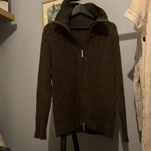 Brun stickad kofta med dragkedja - Säljer en snygg brun stickad kofta med dragkedja. Koftan har en hög krage och långa ärmar, perfekt för kyliga dagar. Den ribbade designen ger en stilren look. Skick 7/10 (inga defekter)