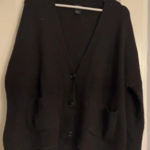 Svart kofta från Lindex - Säljer en stilren svart kofta från Lindex. Den har en v-ringad design med knappar framtill och praktiska fickor. Perfekt för lager-på-lager under kyligare dagar.