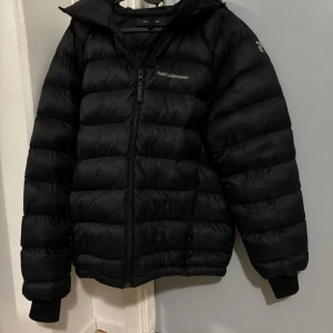 Svart dunjacka från Peak Performance - Säljer en svart dunjacka från Peak Performance. Jackan har en pufferdesign med dragkedja framtill och en justerbar huva. Perfekt för kyliga dagar.