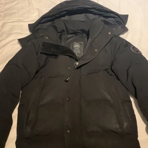 Canada goose wyndham - Säljer en svart dunjacka från Canada Goose med huva och dragkedja. Jackan har långa ärmar och en broderad logga på ärmen. Perfekt för kalla dagar.