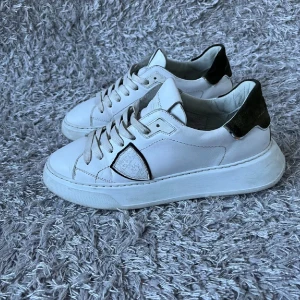 Vita sneakers med svarta detaljer - Snygga vita sneakers med svarta detaljer och snörning. Skorna har en stilren design med en tjock sula och är perfekta för en casual look. De har en svart häl och en diskret logga på sidan. Dessutom kan jag putsa upp de så de bli riktigt fräscha!