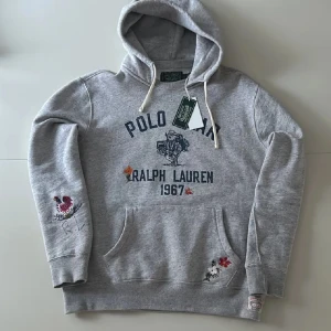 Grå hoodie från Ralph Lauren - Säljer en grå hoodie från Ralph Lauren med broderade detaljer. Tröjan har en stor ficka framtill och en justerbar huva med snören. Perfekt för en avslappnad stil!
