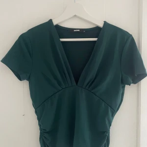 Grön v-ringad topp från Bik Bok - Snygg grön topp från Bik Bok med v-ringning och korta ärmar. Toppen har en smickrande passform med rynkade detaljer i midjan. Perfekt för en stilren look.