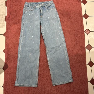 Blå jeans med pärldetaljer - Snygga blå jeans med pärldetaljer på framsidan. De har en klassisk femficksdesign och en vid passform som ger en avslappnad look. Perfekta för att lägga till lite extra stil i vardagsgarderoben.
