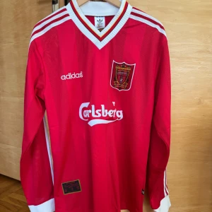 Liverpool matchtröja från Adidas - Klassisk röd fotbollströja från Adidas med vit V-ringning och långa ärmar. Tröjan har klubbmärke och sponsorlogga på framsidan. Perfekt för fotbollsfans som vill ha en retrolook. Storlek L men känns som M.