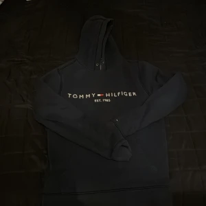 Svart hoodie från Tommy Hilfiger - Säljer en klassisk svart hoodie från Tommy Hilfiger med logotypen tryckt på framsidan. Hoodien har en bekväm passform och är perfekt för en avslappnad stil. Den har långa ärmar och en praktisk huva.