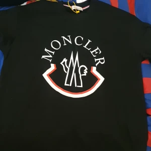 Svart t-shirt från Moncler - Snygg svart t-shirt från Moncler med stort vitt och rött tryck på framsidan. T-shirten har en klassisk rund halsringning och korta ärmar. Perfekt för en stilren look. Storlek M men passar L också. Priset kan diskuteras. 