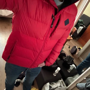 Röd pufferjacka - Säljer en snygg röd pufferjacka med dragkedja och hög krage. Jackan har långa ärmar och praktiska fickor med dragkedja. Perfekt för kyliga dagar. Har en liten lagning.