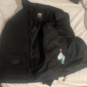 Svart dunjacka från Canada Goose - Säljer en svart dunjacka från Canada Goose med deras ikoniska logga på ärmen. Jackan har en dragkedja och är perfekt för kalla vinterdagar. Den är långärmad och har en klassisk design som håller dig varm och stilren.