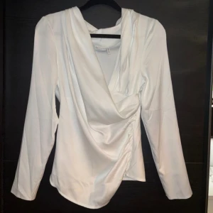 Vit omlottblus - Elegant vit omlottblus med långa ärmar och draperad design. Blusen har en diskret dragkedja på sidan för en smidig passform. Perfekt för en stilren look.