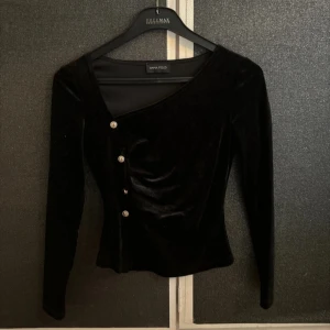Svart sammetstopp från Anna Field - Elegant svart sammetstopp från Anna Field med asymmetrisk design och dekorativa knappar framtill. Toppen har långa ärmar och en figurnära passform, perfekt för en stilren look.