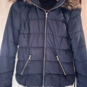 Snygg mörkblå täckjacka från Divided! - Snygg mörkblå figursydd täckjacka med fickor fram och avtagbar luva med fuskpäls i storlek S från Divided. 